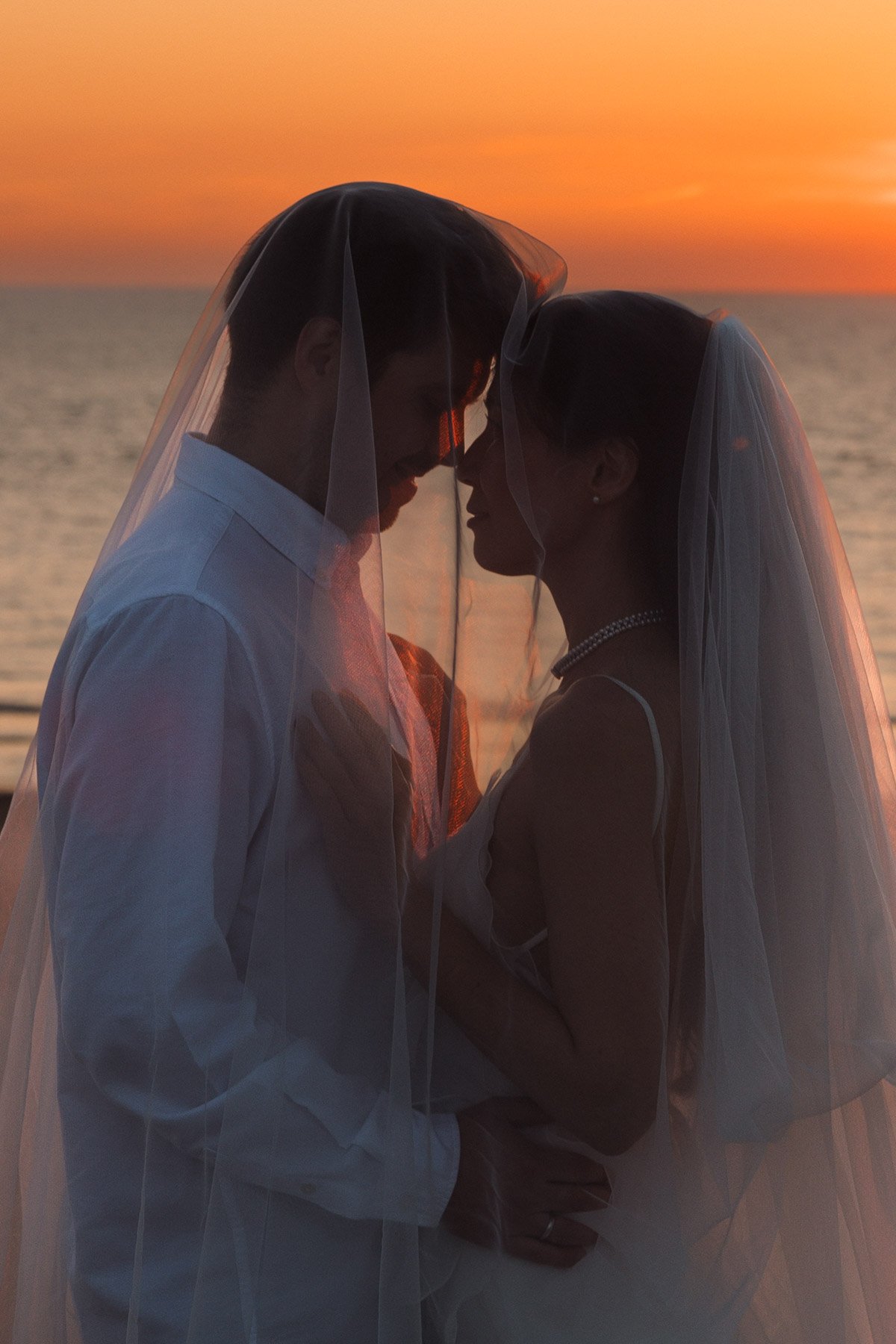 Bride and groom standing beneath a wedding veil at sunset by the sea
